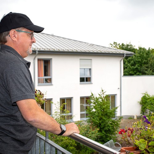 Ein Mann mit einer schwarzen Kappe steht auf einem Balkon mit Grünpflanzen und sieht in einen Garten herunter Ein Mann mit einer schwarzen Kappe steht auf einem Balkon mit Grünpflanzen und sieht in einen Garten herunter